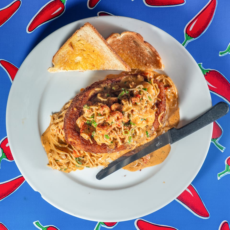 White plate of creamy crawfish pasta topped with a breaded cutlet, served with toasted bread on a red pepper-patterned tablecloth.