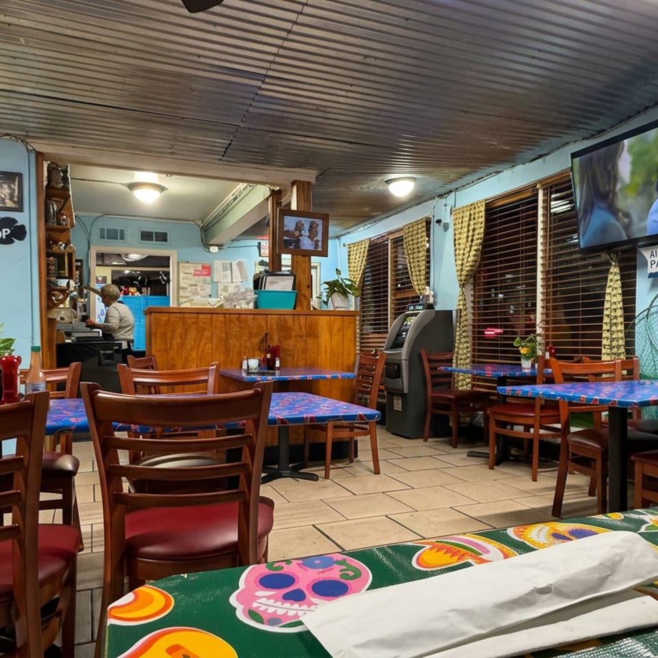 Colorful casual restaurant dining room with wooden tables, patterned tablecloths, yellow doors, television, and decorative curtains.