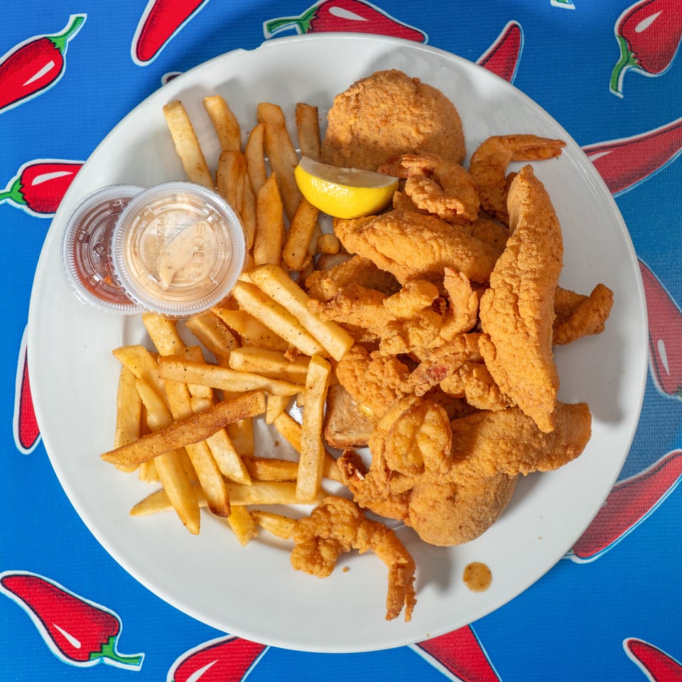 Plate of fried seafood, French fries, dipping sauces, and lemon wedge on a red pepper-patterned blue tablecloth.
