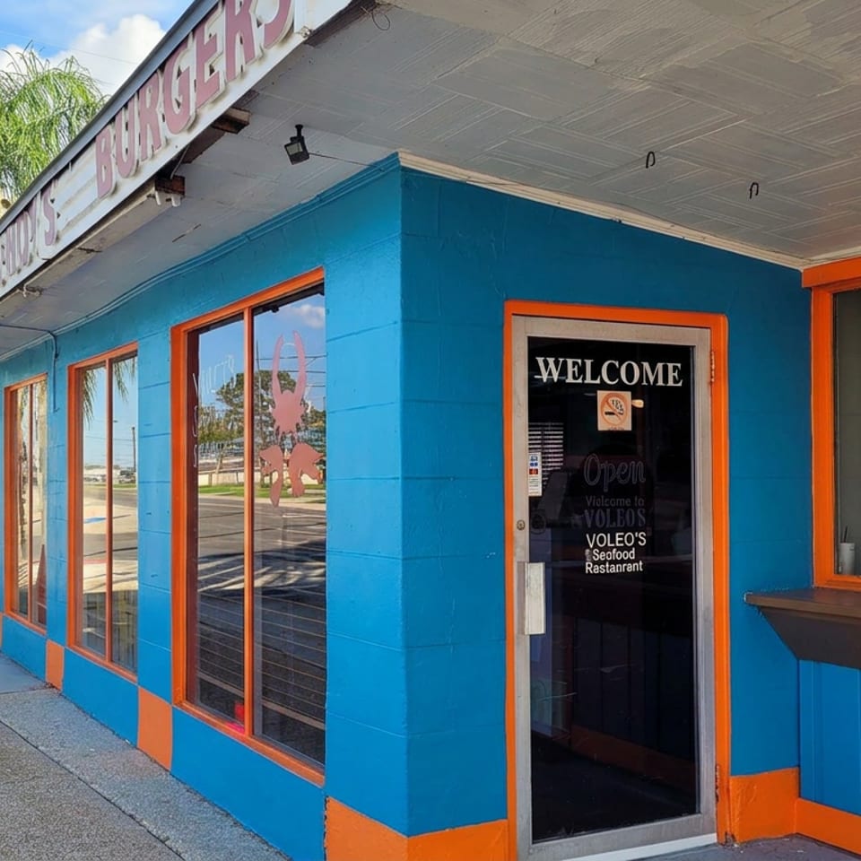 Bright blue and orange Voleo’s Seafood Restaurant storefront with glass entrance, welcome sign, sidewalk, and palm trees.