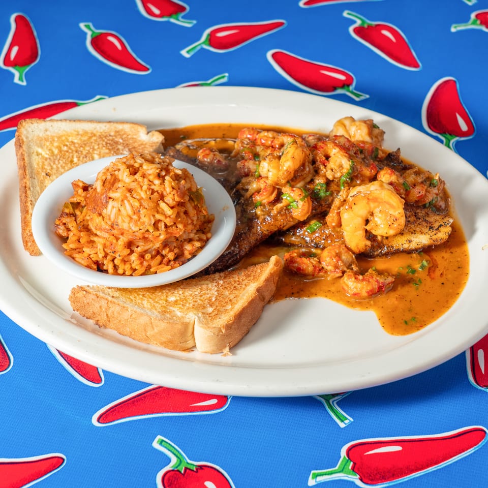 Plate of shrimp-topped grilled fish with seasoned rice and toast in sauce, served on a red chili pepper-patterned tablecloth.