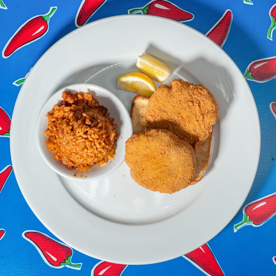 Plate of golden breaded fish, seasoned rice, lemon wedges, and toast on a red chili pepper-patterned tablecloth.