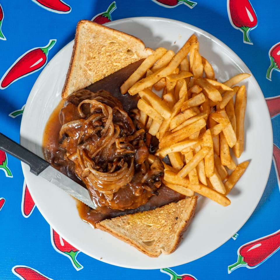 Plate of steak smothered in mushroom gravy with French fries and toasted bread on a red pepper-patterned tablecloth.
